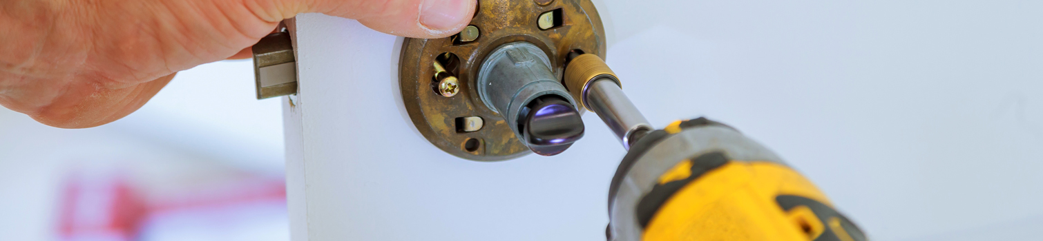 Detalle de la mano de un técnico de keys cerrajeros apretando los tornillos del cilindro de una cerradura con un atornillador eléctrico en una puerta blanca.